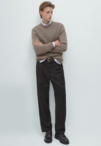 Hnědý pletený svetr, bílá košile s límečkem pod ním, tmavě hnědé kalhoty a tmavé boty. Oblečení má volný střih a texturovanou tkaninu.