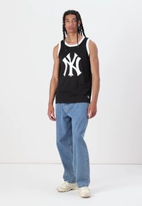 Juodas be rankovių marškinėlis su baltu NYC logotipu, derinamas su šviesiai mėlynomis laisvomis džinsomis ir kreminių spalvų sportinėmis bateliais, pateiktas prieš baltą foną.