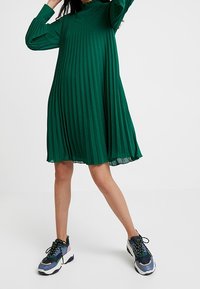 Vihreä laskostettu mekko pitkillä hihoilla ja korkealla kaula-aukolla, yhdistettynä paksuihin, monivärisiin tennareihin. Kevyt kangas, jossa virtaava muoto.