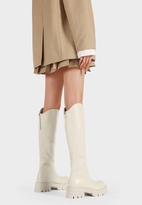 Stivali bianchi al ginocchio realizzati in materiale lucido con suola spessa, dettaglio con zip e scanalature testurizzate sulla suola per una maggiore aderenza.