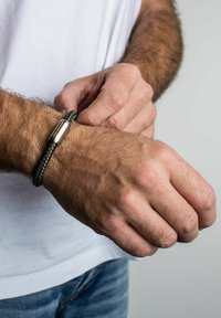 Žalias pintas apyrankė su metaliniu užsegimu, dėvima ant riešo. Dizainas pasižymi tekstūruotu pynimu ir lygiu, blizgiu akcentu.
