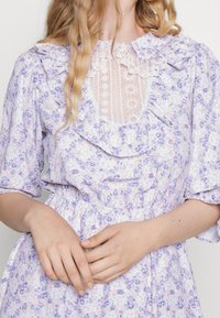 Žena na sobě má levandulové květinové šaty s nabíranými rukávy, volánkovým límcem a průhlednou krajkovou panelou na hrudi, ruce spojené v pase.