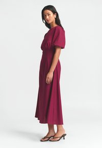 Dolga burgundy obleka z napihnjenimi rokavi in prilegajočim se korzetom, ki prehaja v široko, dolgoprednjo krilo do gležnjev. V paru z črnimi natikači.