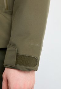 Manche d'une veste vert olive avec une sangle réglable en Velcro et un logo "DRYVENT" discret au-dessus du poignet sur le bras d'une personne.