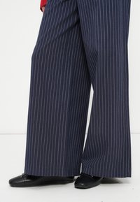 Pantalons larges bleu marine à rayures, avec une finition texturée, associés à des chaussures plates noires ornées d'un accent décoratif sur l'orteil.
