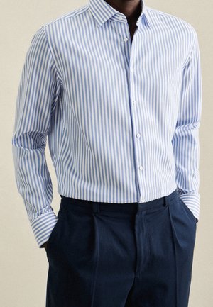 Man draagt een slim-fit, lange mouwen, blauw-wit verticaal gestreept overhemd met knopen, ingestopt in donkerblauwe broek.
