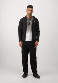 Vīrietis ar melnu kapuci, melnām kravas biksēm un melnām sporta apaviem, stāvot uz vienkārša gaiša fona.