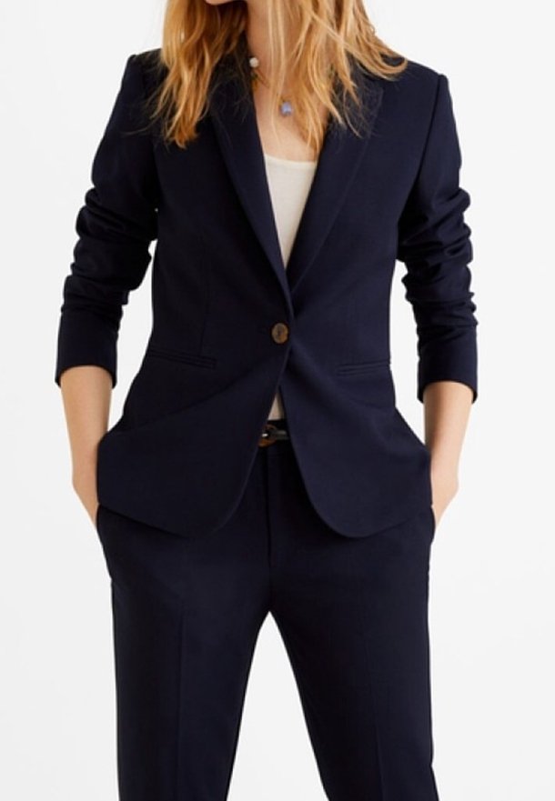 Femme portant un blazer bleu marine ajusté avec un bouton, un haut blanc et un pantalon bleu marine assorti, mains dans les poches, sur un fond uni.