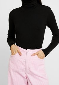 Pull noir à col roulé avec texture côtelée, associé à un pantalon en velours côtelé rose taille haute avec poches latérales et fermeture par boutons.