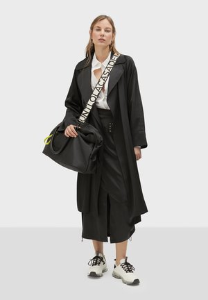 Abrigo trench oversized negro con solapas anchas, camisa blanca, falda negra con aberturas, bolso negro con acentos neón y zapatillas blancas gruesas.