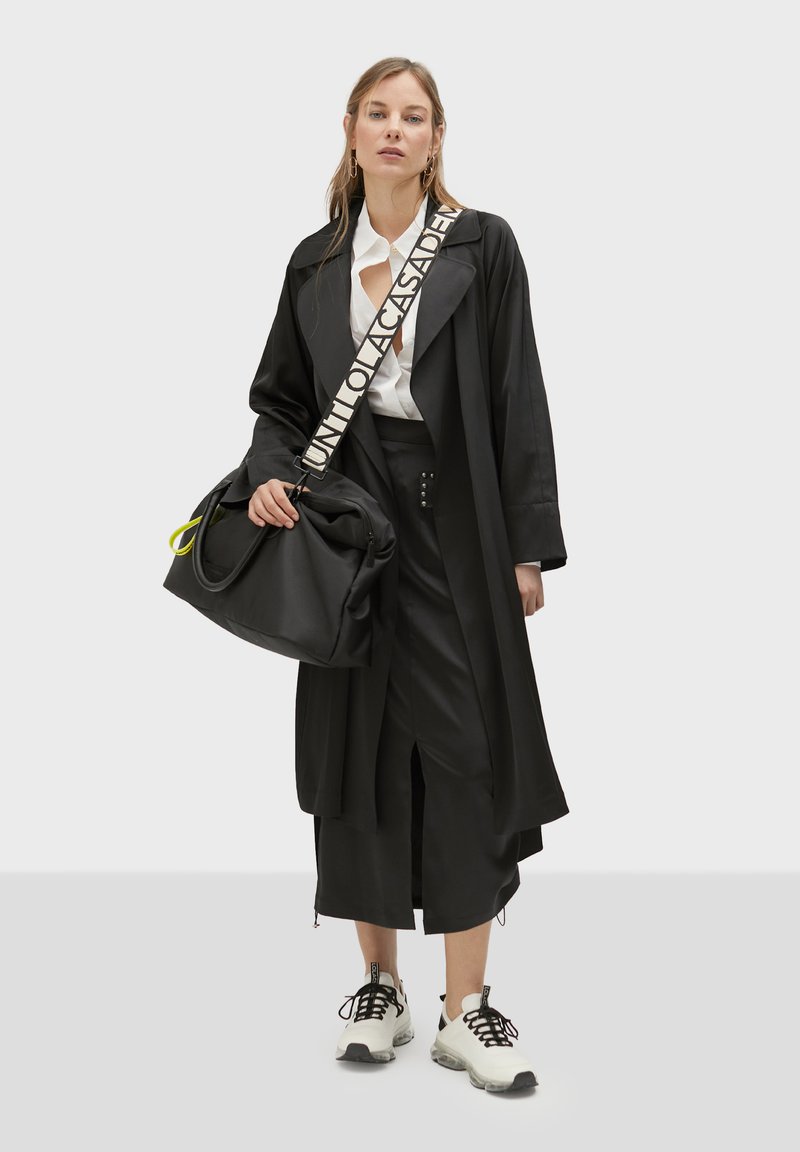 Abrigo trench oversized negro con solapas anchas, camisa blanca, falda negra con aberturas, bolso negro con acentos neón y zapatillas blancas gruesas.
