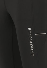 Zwarte leggings van rekbaar materiaal, met een wit "ENDURANCE" logo, een klein zakje en contraststiksels langs de naden.