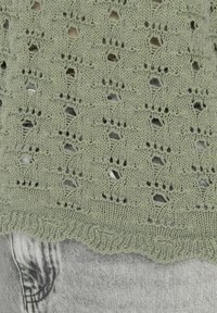 Suéter de punto en verde oliva claro con diseño de calado que presenta formas de diamante. Superficie texturizada y dobladillo ondulado sobre unos vaqueros de mezclilla grises.