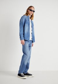 Giacca di jeans blu chiaro con due tasche sul petto, abbinata a jeans coordinati. Sneakers nere con accenti bianchi. Silhouette slim e dritta.