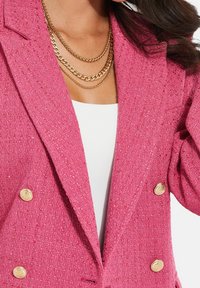 Veste rose texturée avec boutons dorés, revers proéminents, et colliers à chaînes dorées superposées sur un haut blanc.