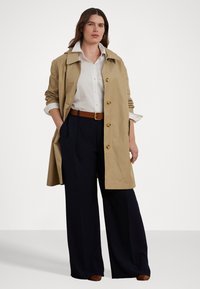 Beige trenchcoat med en avslappnad passform, stor krage och guldknappar, matchad med en vit skjorta och marinblå vida byxor.
