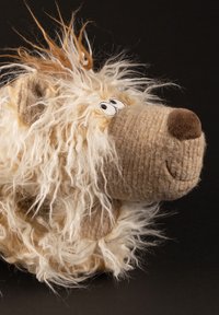 Gestrickte beige Bärenpuppe mit flauschiger weißer und orangefarbener Wolle als Mähne, gestickten Augen, lächelndem Mund und großer brauner Nase vor schwarzem Hintergrund.