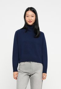 Giovane donna con capelli lunghi neri, vestita con un maglione a collo alto blu navy e jeans grigio chiaro, in piedi davanti a uno sfondo bianco.