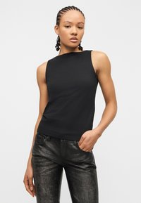 Femme aux cheveux tressés portant un haut noir sans manches ajusté et un pantalon en cuir noir texturé, debout avec une main dans la poche.