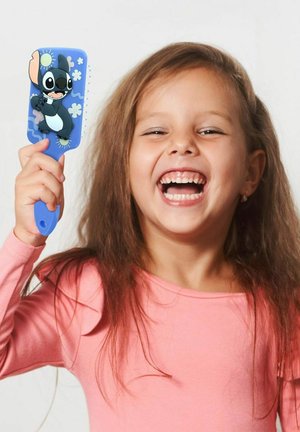 Ein lächelndes junges Mädchen mit hellbraunen Haaren hält eine blaue Haarbürste mit einem Cartoon-Charakter und trägt ein pinkes Langarmshirt.