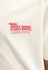 Biely bavlnený tričká s červeným potlačeným textom: "Servis plaváckych opravovní Superior Pool Repair," s údajmi o mieste nižšie hlavného textu.