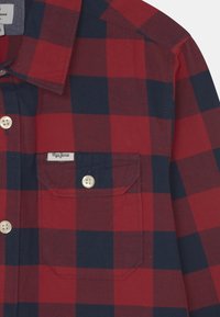 Czerwono-granatowa koszula flanelowa w kratę z kołnierzykiem zapinanym na guziki, kieszenią na piersi i metką nad kieszenią. Bawełniana faktura.