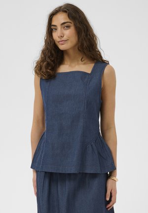 Femme aux cheveux bouclés portant un haut sans manches en denim bleu foncé avec un décolleté carré et une jupe assortie, sur fond uni.