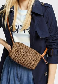 Brązowa, pleciona torba na ramię wykonana z materiału przypominającego słomę, z gładkim, jasnobrązowym skórzanym paskiem i prostokątnym kształtem.
