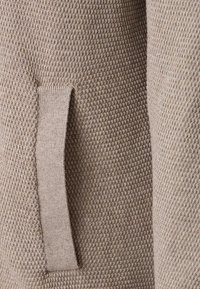 Beige strukturerad tyg med ett bikakesmönster och en sidficka. Tyget har en mjuk, stickad känsla och subtila tonala variationer.