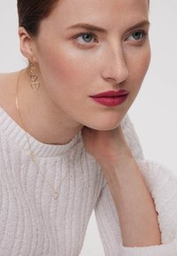 Złote kolczyki w kształcie serca oraz pasujący delikatny złoty naszyjnik na modelce w białym, fakturowanym swetrze.