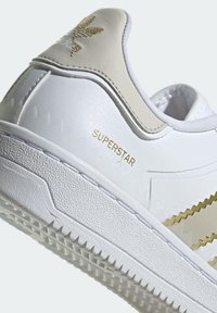 Bílé kožené tenisky s texturovaným povrchem. Obsahují béžový akcent, zlatý nápis "Superstar" a zubatý design po stranách.