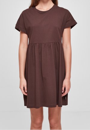 Vrouw in een kortere, loszittende bruine jurk met een gerimpelde taille en een ronde hals, staand tegen een effen achtergrond.