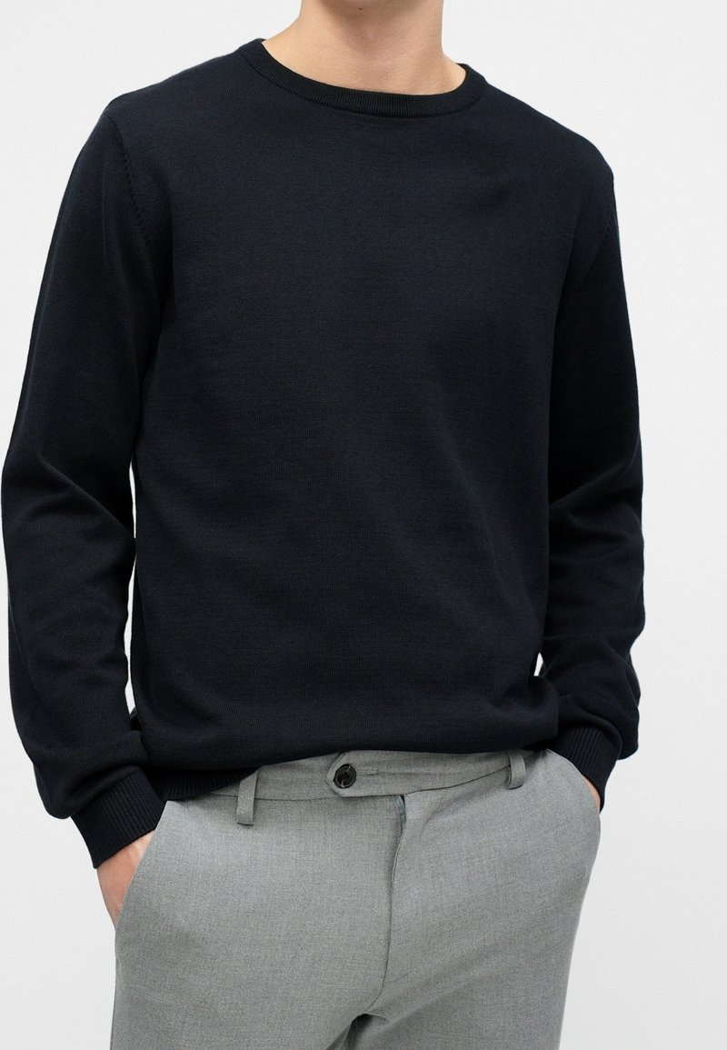 Personne portant un pull noir uni à manches longues et un pantalon gris clair, les mains dans les poches, sur un fond neutre.