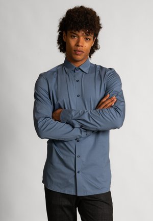 Jonge man met krullend haar, die een blauw overhemd met lange mouwen en knopen en een zwarte broek draagt, staat met gekruiste armen tegen een effen achtergrond.