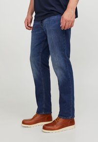 Tummansinisiä farkkuja suoranlahkeisella leikkauksella, joissa on kulutettuja yksityiskohtia ja kontrastiväristä ommelta; yhdistettynä ruskeisiin nahkabootseihin, joissa on keltaiset nauhat.