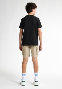 T-shirt in cotone nero indossata con pantaloni cargo beige. Calze bianche con strisce blu e sneaker bianchi con dettagli verdi. Vista da dietro.