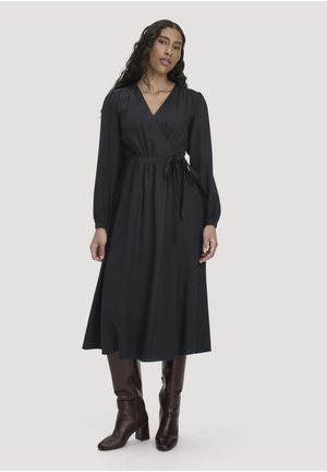 Femme debout portant une robe portefeuille noire à manches longues avec une ceinture à la taille et des bottes hautes marron foncé sur un fond clair.