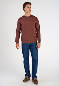 Brun långärmad sweatshirt med ribbad textur, med rundad halsringning och subtil logotypdetalj. Bärs med blå denimjeans och bruna skor.