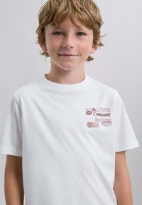 Camiseta de algodón blanca con texto impreso en rojo en el lado izquierdo del pecho que dice "SCLPERS The Company" con detalles adicionales debajo. Mangas cortas.