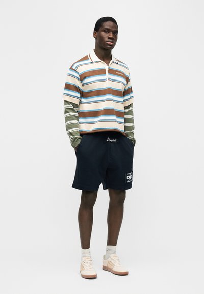 Jeune homme portant un polo rayé marron et bleu en couches avec des manches longues rayées vertes, un short bleu marine, des chaussettes blanches et des baskets beiges, debout.