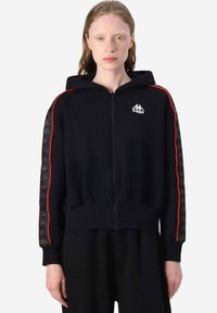 Felpa nera con zip, strisce laterali rosse, logo Kappa sul petto, maniche nere con motivo testurizzato, polsini e orlo a costine. Tessuto morbido.