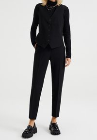 Svart väst med fem knappar, i kombination med avsmalnande svarta byxor. För att fullända looken finns blanka svarta loafers med tjock sula.