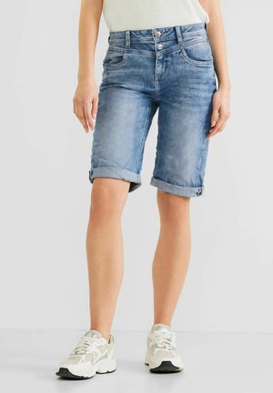 Person trägt hochgekrempelte hellblaue Jeansshorts und weiße Turnschuhe und steht vor einem einfarbigen Hintergrund.