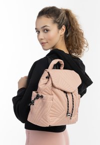 Pudret pink quiltet rygsæk med klaplukning, sorte snore og tekstureret design. Bæres over en sort hoodie.