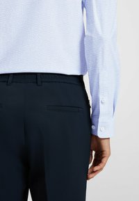 Vaaleansininen paita, jossa on teksturoidun näköinen kuvio, yhdistettynä tummansinisiin housuihin, joissa on joustava vyötär ö ja takataskun yksityiskohta.