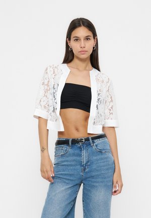 Ung kvinde iført en hvid blonder cropped jakke over en sort bandeau top og blå højttaljede jeans med sort bælte, stående mod hvid baggrund.