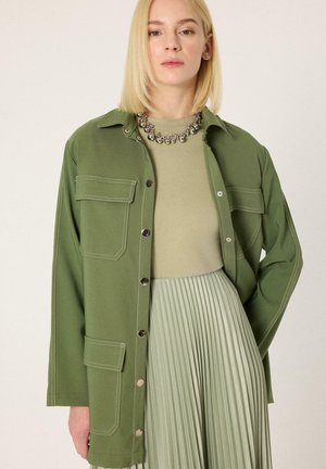 Lyshåret kvinde iført en olivengrøn jakke med knapper, lysegrøn top og plisseret lysegrøn nederdel, accessoriseret med en perlekæde.