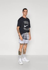 Melna T-krekls ar baltu grafisku zīmējumu un Nike logotipu, rakstainas pelēkās un baltās krāsās šorti, baltas sporta kurpes un potītes zeķes.