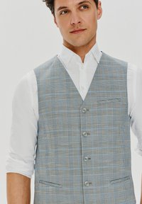 Szary kamizelka w kratę z niebieskimi akcentami, charakteryzująca się klasycznym krojem, pięcioma guzikami i dwiema przednimi kieszeniami, noszona na białej koszuli.