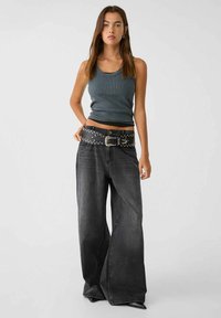 Top tank grigio a righe abbinato a jeans neri in denim a gamba larga. La cintura è decorata con borchie e una fibbia di metallo, aggiungendo un tocco di stile all'outfit.